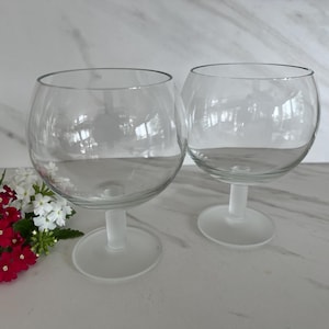 May include: Two clear, round-bowled wine glasses with frosted stems and bases. The glasses are empty and sit on a white surface. A small bouquet of red and white flowers is in the lower left corner.