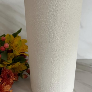 May include: A tall, off-white textured vase. The cylindrical vase has a rough, stucco-like surface. A bouquet of yellow, orange, and red flowers is partially visible to the left of the vase. The vase is likely for home decor.