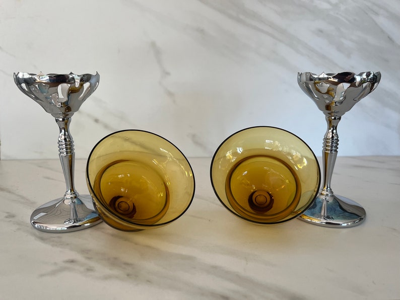 May include: Two champagne glasses with amber-colored bowls and silver-colored stems. The bowls are tilted, revealing their interior design. The stems have a decorative, ribbed design, and the bases are round.