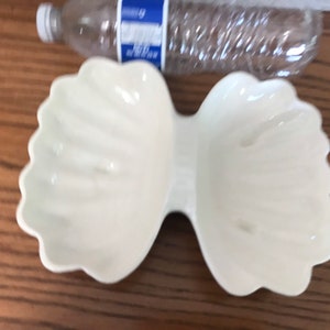 May include: A white, bow-shaped ceramic dish with scalloped edges. The dish has two separate compartments connected in the center. A clear plastic water bottle is visible in the background.