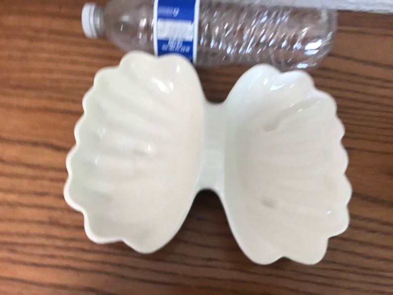 May include: A white, bow-shaped ceramic dish with scalloped edges. The dish has two separate compartments connected in the center. A clear plastic water bottle is visible in the background.