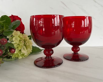 May include: Two red wine glasses with a stemmed base. The glasses are made of clear glass and have a deep red color.