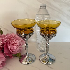 May include: Two amber-colored glass candle holders with a silver-colored metal base and stem. The candle holders have a wide, shallow bowl shape. A clear plastic water bottle is in the background, along with pink flowers.