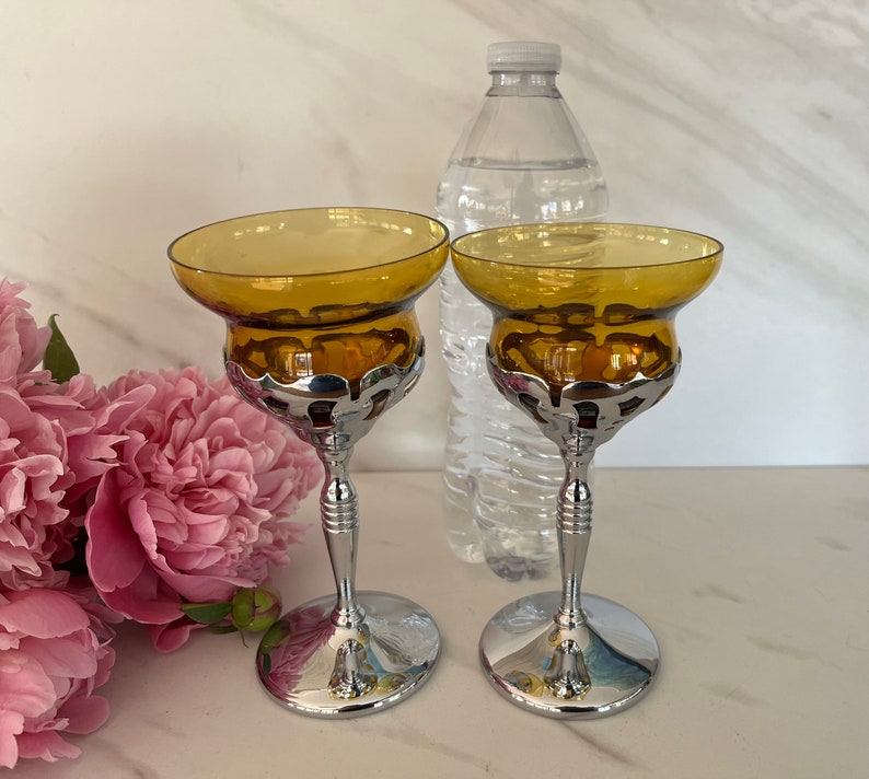 May include: Two amber-colored glass candle holders with a silver-colored metal base and stem. The candle holders have a wide, shallow bowl shape. A clear plastic water bottle is in the background, along with pink flowers.