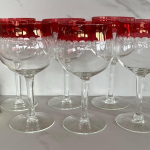 May include: A set of clear wine glasses with a red scalloped rim. The glasses have a long stem and a round base. The image shows a close-up of the glasses, with a few roses in the background. The glasses are likely for a special occasion.
