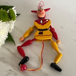 May include: A colorful wooden marionette doll with a clown-like face, red horns, and a yellow outfit. The doll's arms and legs are painted in red, yellow, and black. A red string is attached for manipulation. White flowers and green leaves are in the background.