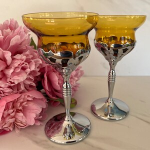 May include: Two elegant, vintage-style goblets with amber-colored bowls and ornate silver-toned stems and bases. The goblets have a unique, decorative design around the bowl. Pink peonies are in the background.
