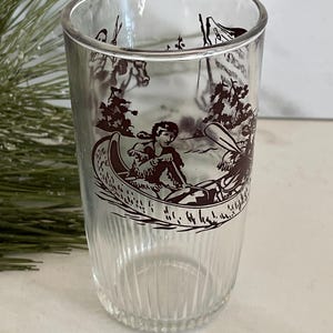 Peut inclure: Verre transparent avec une illustration marron d'une personne en canoë. Le verre présente des détails verticaux côtelés. L'illustration représente une personne pagayant en canoë sur un lac ou une rivière, entourée d'arbres et d'autres personnages.