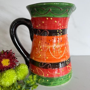 May include: A hand-painted ceramic pitcher with a black handle. The pitcher has a green, red, orange, and black striped design with yellow accents.