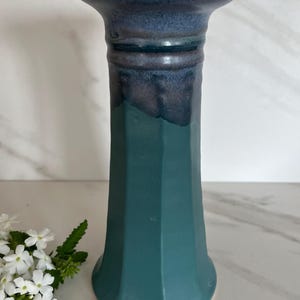 May include: A ceramic vase with a teal and dark blue glaze. The vase has a wide, flared top and a faceted body. The top is dark blue, transitioning to teal. White flowers are in the background.