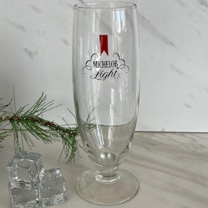 May include: A clear glass beer pilsner with a round base and a tall, slender body. The glass features the "Michelob Light" logo in black script with a red accent. Ice cubes and a sprig of greenery are in the foreground.