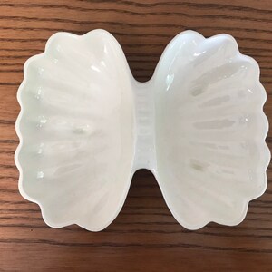 May include: A white, bow-shaped ceramic dish with scalloped edges. The dish has a glossy finish and is designed with two distinct sections connected by a narrow center. The dish is sitting on a wooden surface.