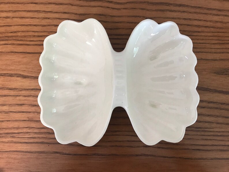May include: A white, bow-shaped ceramic dish with scalloped edges. The dish has a glossy finish and is designed with two distinct sections connected by a narrow center. The dish is sitting on a wooden surface.
