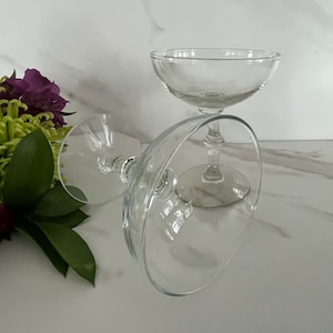 May include: Two clear glass champagne coupes stacked on top of each other. The glasses have a wide bowl and a long, thin stem.
