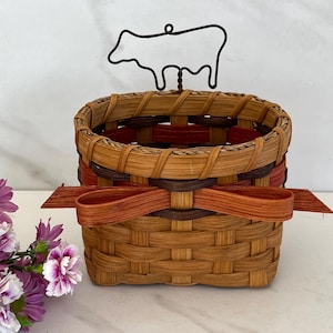 May include: A woven basket with a rectangular shape, featuring a brown and reddish-brown color scheme. A decorative bow is attached to the front. A black metal cow silhouette is attached to the top of the basket. The basket is likely used for storage or decoration.
