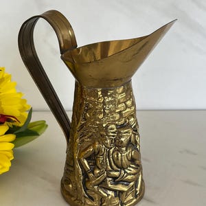 May include: A gold brass pitcher with an embossed design of people and a building. The pitcher has a wide spout and a curved handle.