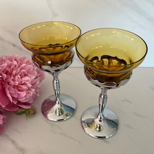 May include: Two vintage champagne coupe glasses with amber-colored bowls and chrome-plated stems. The glasses have a wide, shallow bowl design and are set on a reflective surface. A bouquet of pink peonies is in the background.