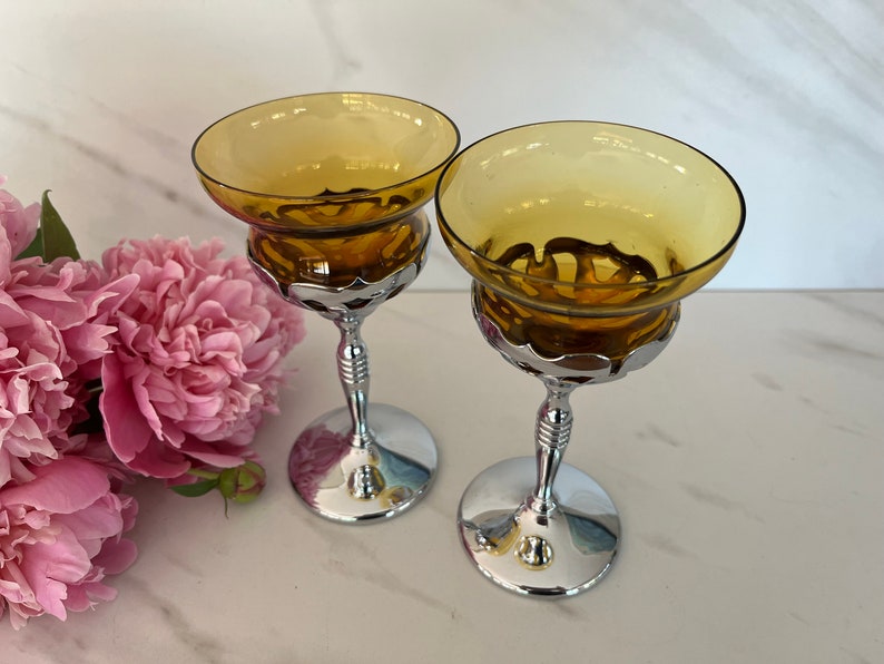 May include: Two vintage champagne coupe glasses with amber-colored bowls and chrome-plated stems. The glasses have a wide, shallow bowl design and are set on a reflective surface. A bouquet of pink peonies is in the background.