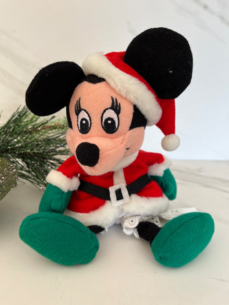 Puede incluir: Un juguete de peluche de Mickey Mouse vestido de Pap&aacute; Noel. El juguete lleva un traje de Pap&aacute; Noel rojo y blanco con un gorro y guantes verdes. El juguete est&aacute; sentado sobre una superficie blanca.