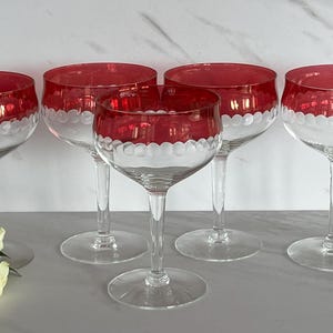 May include: Five clear champagne glasses with red rims and a white scalloped design. The glasses are arranged on a light-colored surface, with a bouquet of red and white roses in the foreground. The glasses have a classic, elegant design.