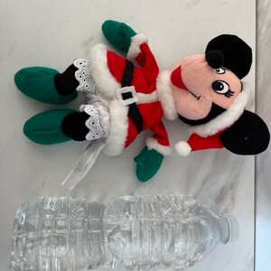 Puede incluir: Un juguete de peluche de Minnie Mouse vestida de Pap&aacute; Noel rojo y blanco con guantes verdes y zapatos negros. El juguete est&aacute; tumbado sobre una superficie blanca junto a una botella de agua de pl&aacute;stico transparente.