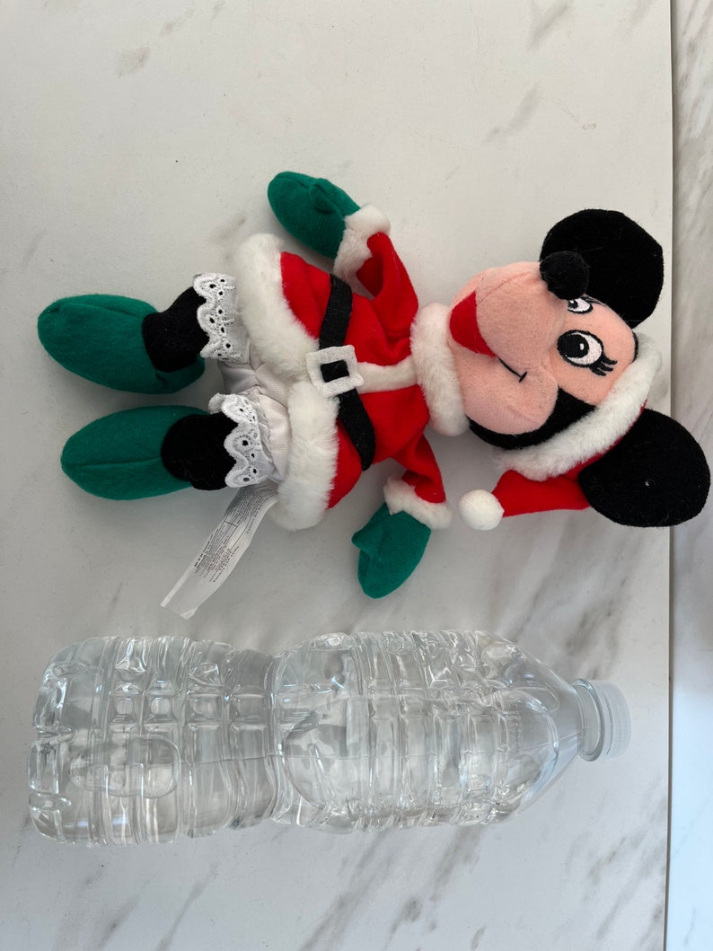 Puede incluir: Un juguete de peluche de Minnie Mouse vestida de Pap&aacute; Noel rojo y blanco con guantes verdes y zapatos negros. El juguete est&aacute; tumbado sobre una superficie blanca junto a una botella de agua de pl&aacute;stico transparente.