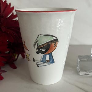 May include: A white ceramic tumbler featuring a hand-painted illustration of a character wearing a green cap, a black mask, and a blue jacket. The cup has a thin red rim and a textured surface, showcasing a whimsical cartoon figure in profile.