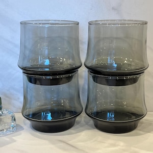 May include: Four gray-tinted glass tumblers stacked in pairs. Each tumbler has a rounded base and a slightly flared rim. The glasses are designed with a modern aesthetic, suitable for serving beverages.