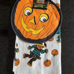 May include: A round orange pumpkin pot holder with a black border and a tag reading Totally Ghoul, paired with a white kitchen towel featuring a repeating pattern of orange pumpkins and scarecrows wearing black and teal checkered shirts.