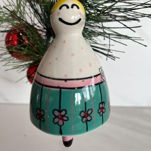 May include: A ceramic Christmas bell ornament with a smiling face and blonde hair. The ornament features a white body with pink polka dots, a pink band, and a green skirt with black stripes and flower designs. A gold string is attached for hanging.