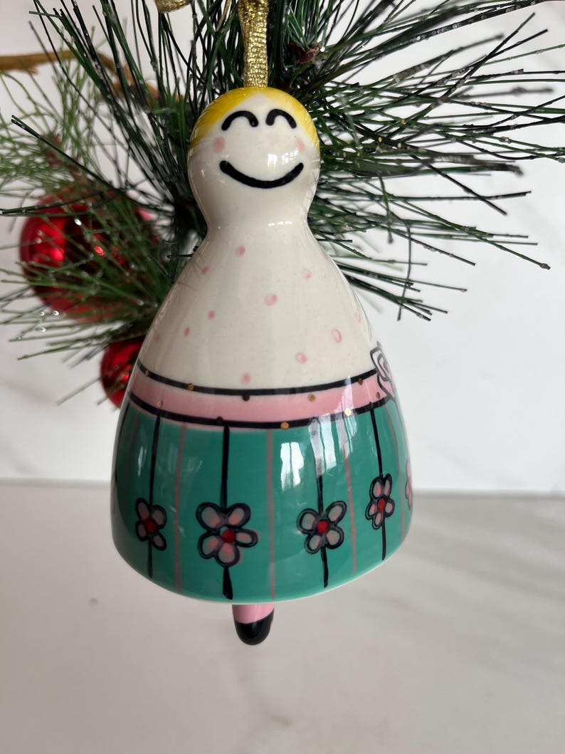 May include: A ceramic Christmas bell ornament with a smiling face and blonde hair. The ornament features a white body with pink polka dots, a pink band, and a green skirt with black stripes and flower designs. A gold string is attached for hanging.