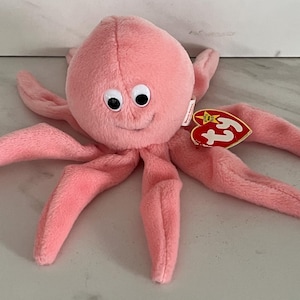 May include: A plush pink octopus stuffed animal with large white and black googly eyes and a stitched smile. The toy features eight soft tentacles and has a red heart-shaped Ty Beanie Baby tag attached to the side, along with a small white fabric label.