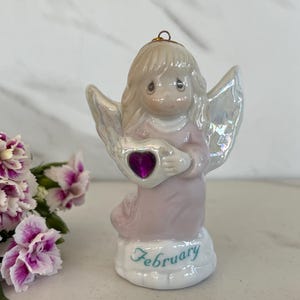 May include: A ceramic angel figurine in soft pink and white tones, holding a heart with a purple gem. The figurine has iridescent wings and is perched on a base with the word "February" in teal script. A bouquet of pink and white flowers is in the foreground.