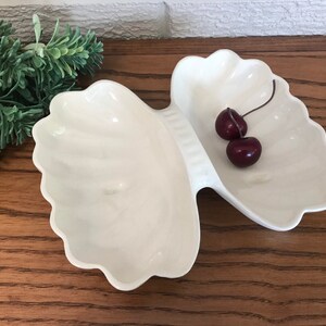 May include: A white, shell-shaped serving dish with two compartments. The dish has a scalloped edge and is filled with two dark red cherries. The dish is on a wooden surface.
