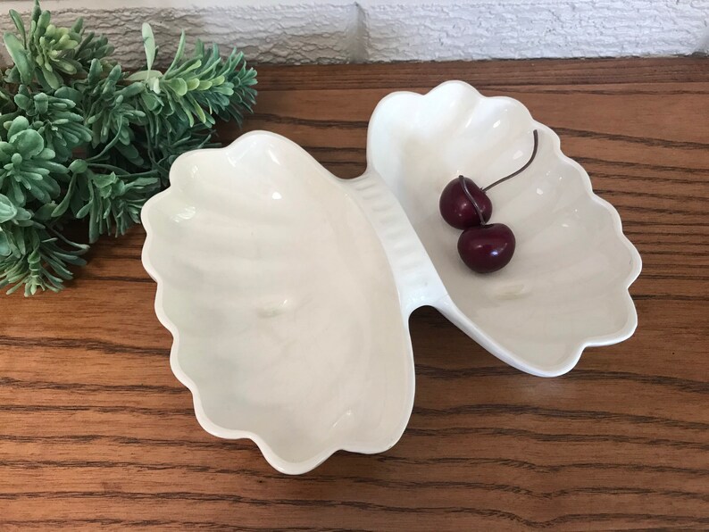May include: A white, shell-shaped serving dish with two compartments. The dish has a scalloped edge and is filled with two dark red cherries. The dish is on a wooden surface.