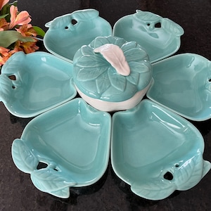 May include: A teal blue ceramic serving dish shaped like a flower with six individual bowls and a central bowl with a lid. The bowls and lid have leaf designs.