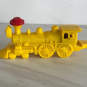 May include: A yellow toy train with a red smokestack. The train has multiple wheels and a detailed design, including a cabin and a front cowcatcher. The words "BUFFALO" and "WOOD" are printed on the side of the cabin.