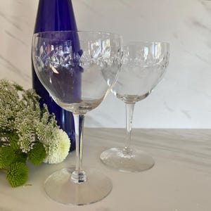 Peut inclure: Deux verres à vin en cristal transparent avec des motifs floraux gravés. Une bouteille bleu foncé est à l'arrière-plan, et un bouquet de fleurs blanches et vertes est à gauche. Les verres sont sur une surface claire.