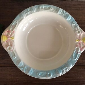 May include: A white ceramic bowl with a blue and pink rim. The rim is decorated with a pattern of baby items, including pacifiers, rattles, and a ribbon. The bowl is perfect for serving baby food or snacks.