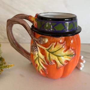 May include: A ceramic mug shaped like a pumpkin, with a brown branch-like handle. The mug is orange with a black and green hat, and features a leaf and acorn design. The mug is a decorative item.