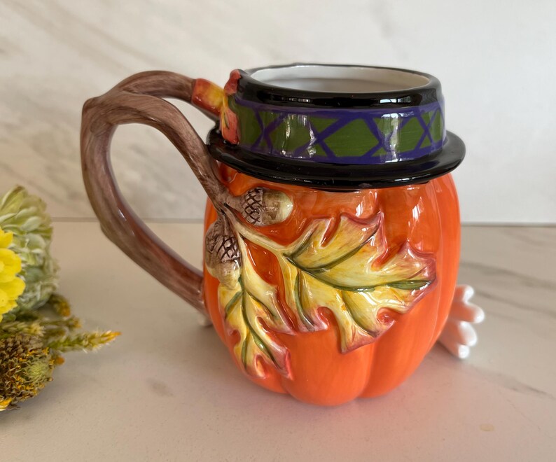 May include: A ceramic mug shaped like a pumpkin, with a brown branch-like handle. The mug is orange with a black and green hat, and features a leaf and acorn design. The mug is a decorative item.
