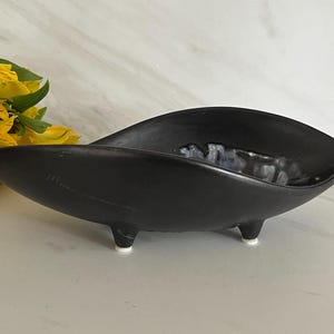 May include: A black ceramic dish with a unique, elongated shape, resting on three small, white-tipped feet. The dish has a textured interior with a marbled, gray and white design. Yellow flowers are visible in the background.
