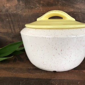 May include: A white ceramic sugar bowl with a yellow lid and a speckled glaze. The lid has a handle.