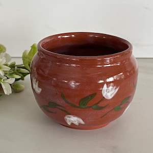 May include: A round, terracotta-colored ceramic bowl with a smooth, glossy finish. The exterior features a hand-painted pattern of small white flowers with green stems and leaves encircling the body of the bowl, which has a wide opening.