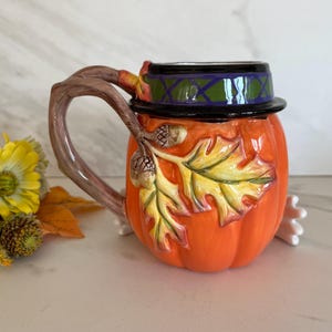 May include: A ceramic pumpkin-shaped mug with a lid, featuring a branch-shaped handle. The mug is orange with autumn leaf and acorn details. The lid is black with a green and purple design. A festive fall-themed mug.