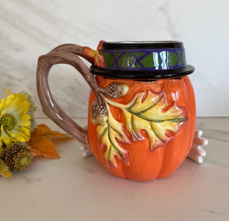 May include: A ceramic pumpkin-shaped mug with a lid, featuring a branch-shaped handle. The mug is orange with autumn leaf and acorn details. The lid is black with a green and purple design. A festive fall-themed mug.