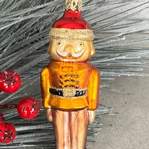May include: A glass Christmas ornament shaped like a nutcracker. The ornament is gold with a red hat and black boots. The nutcracker has a white mustache and a black belt.