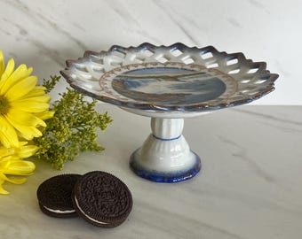 May include: A white and blue ceramic pedestal cake stand with a decorative openwork rim. The center of the plate features a landscape scene. Two chocolate sandwich cookies are in the foreground, with a yellow flower and greenery to the left.