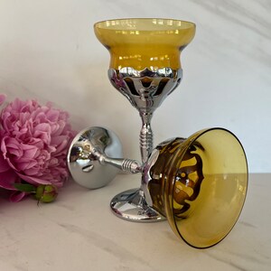 May include: Two decorative goblets with amber-colored bowls and ornate silver-colored stems. One goblet is upright, while the other is lying on its side. The goblets are set against a white background with pink flowers.