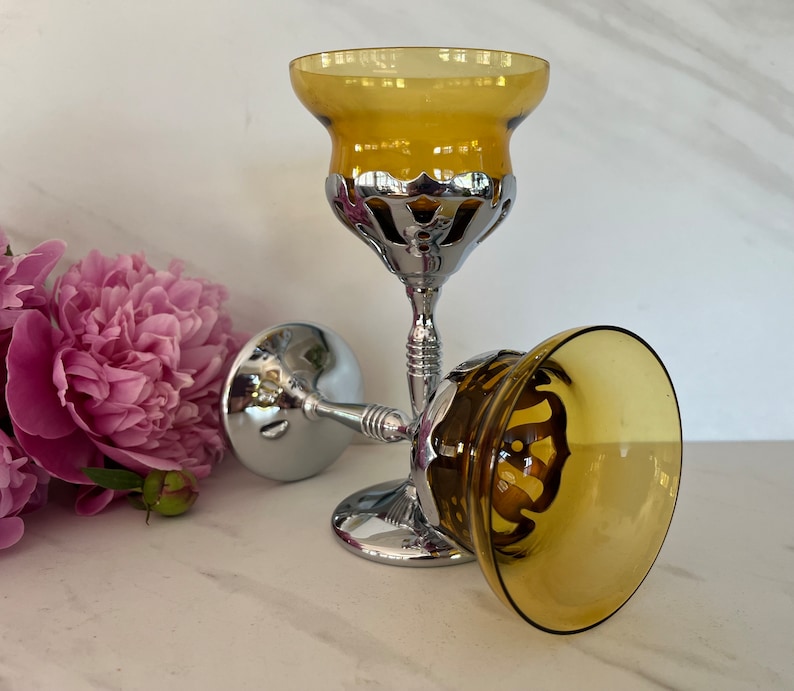 May include: Two decorative goblets with amber-colored bowls and ornate silver-colored stems. One goblet is upright, while the other is lying on its side. The goblets are set against a white background with pink flowers.